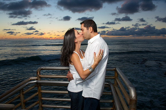 The engagement couple kisses high above the ocean as a lovely rainbow colored sunset appears in the sky behind them at Sunset Cliffs Natural Park in San Diego, CA
