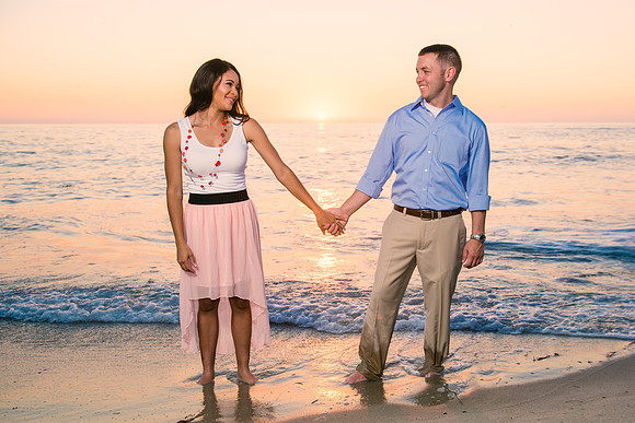 A newly engaged couple standing with their feet in the sand on the beach at Cuvier Park in La Jolla, California, looking at each other, holding hands and smiling with a beautiful multicolored sunset b