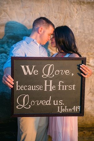 Engagement couple touch foreheads while they hold up a hand lettered chalk sign with a bible verse on it, as they stand at the base of a sandstone beach cliff at sunset