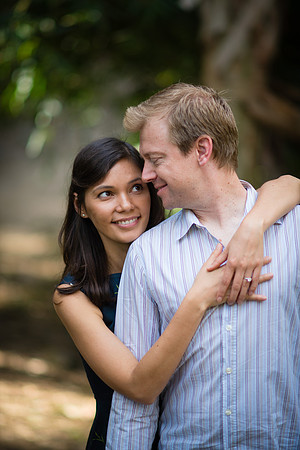 The cute bride puts her arms around her groom to be in a peaceful corner of Balboa Park surrounded by luscious greenery and dappled sunlight