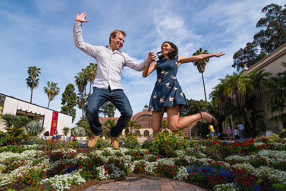 Excited, happy couple jumping into the air while holding hands in front of the Botanic Gardens at Balboa Park in San Diego on a beautiful sunny day.