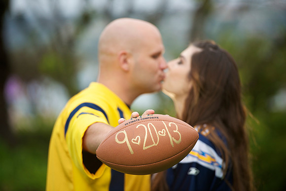 Engagement couple kissing out of focus on a green background while the groom holds up a football with their wedding date written on it