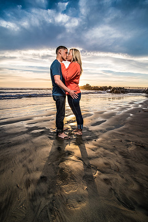 Engagement couple kissing while standing on an empty Coronado beach during a colorful sunset with lots of fluffy clouds visible