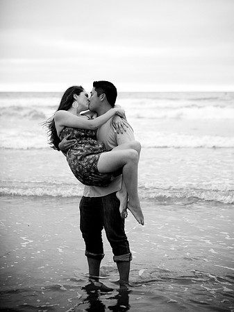 Black and white scene of boy holding girl in his arms with a calm beach in the background near Mission Beach, CA.