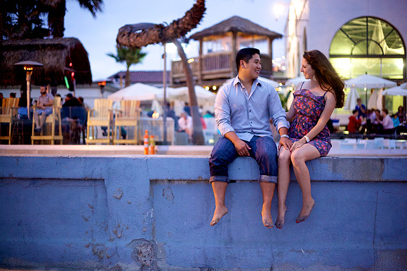 The cute engagement couple sits on a concrete wall at dusk, holding hands as the lights of Mission Bay's Belmont Amusement Park shine behind them
