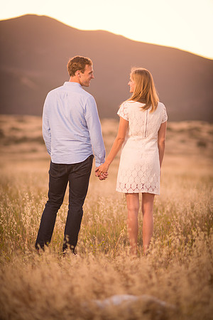 A cute engagement couple stands in a field of grass holding hands and loking at one another, illuminated golden by the sun setting over the mountains