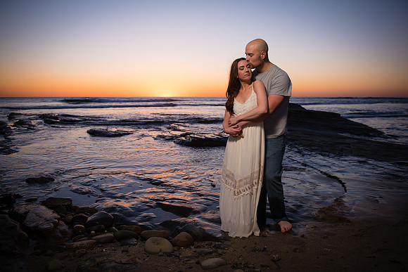 The engagement couple stands with their arms around one another as the multicolored sunset fills the sky and reflects ofd the water's surface above the tide pools at Sunset Cliffs near Ocean Beach, Ca